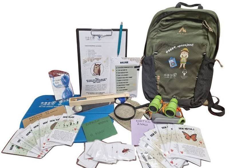 Fotografija nahrbtnika in njegove vsebine med katero so lupa, daljnogled, različni kartončki, lesena puščica, vrvica, pisalo in posalna podloga s papirjem.