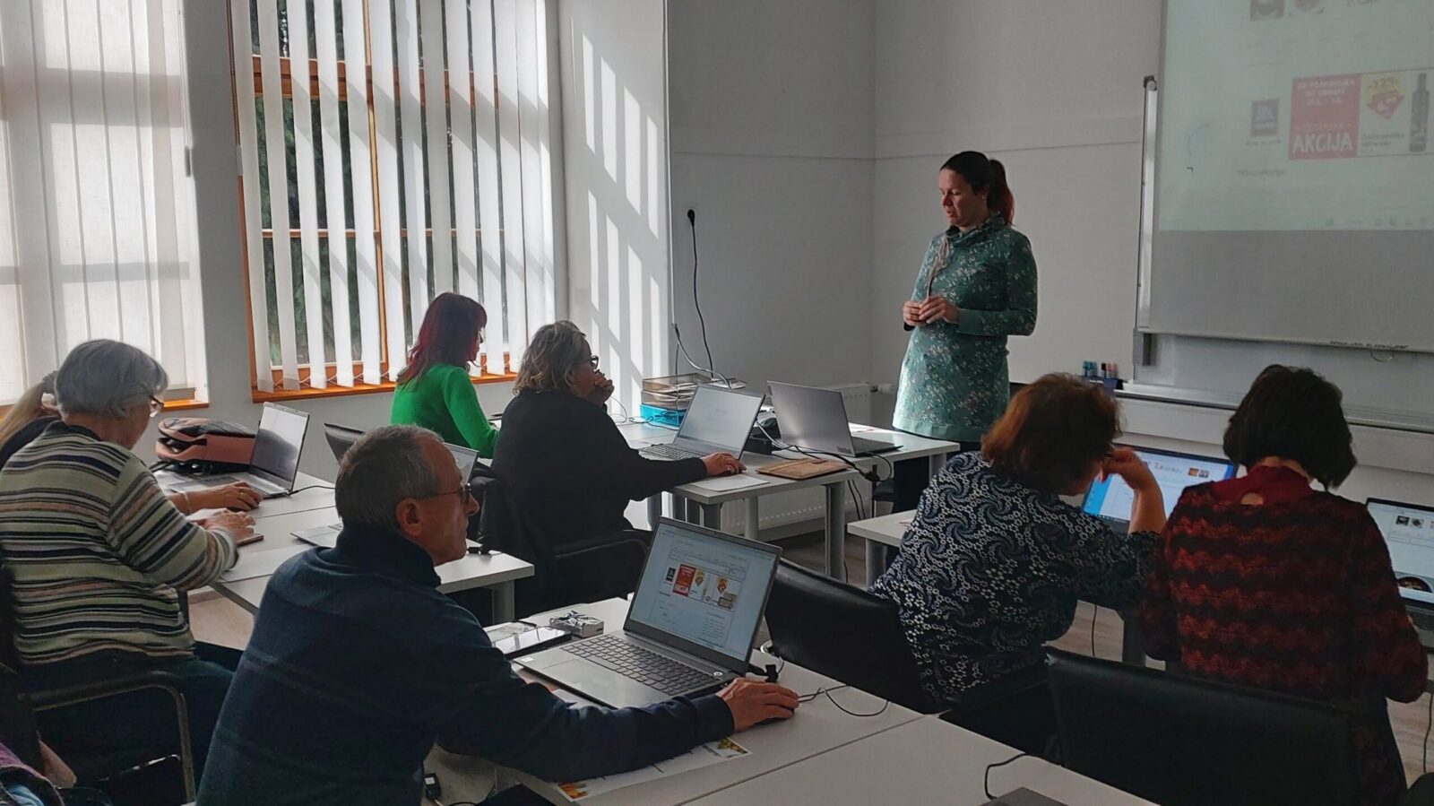 Fotografija z delavnice, udeleženci v učilnici, pred seboj imajo prenosne računalnike, predavateljica stoji pred jimi.