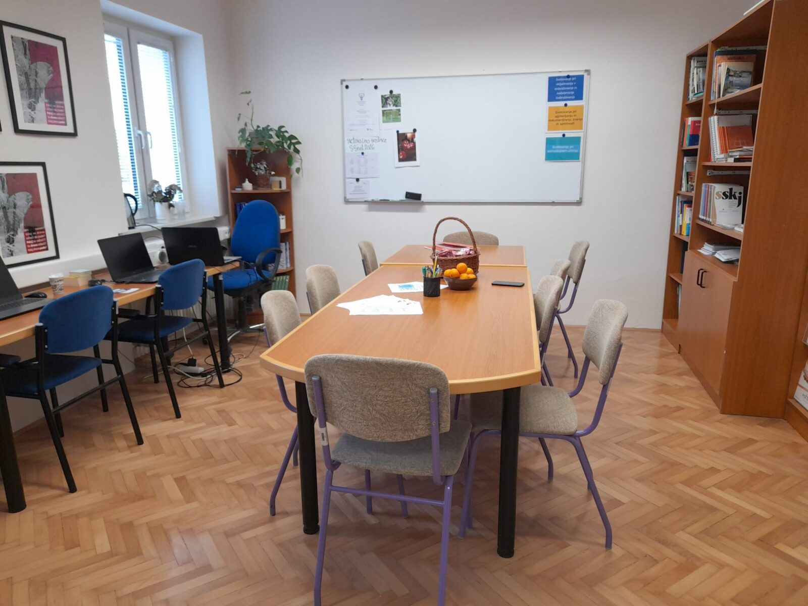 Fotografija SSU na Ljudski univerzi v Murski Soboti. Učna mesta, opremljena z računalniki, velika miza s stoli na sredi sobe in knjižne police.